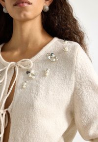 Cardigan beige lavorato a maglia con chiusura a fiocco, decorato con perle e dettagli in argento su spalle e scollo. Tessuto strutturato.