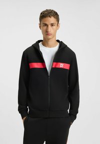 Felpa nera con zip realizzata in tessuto liscio, caratterizzata da una striscia orizzontale rossa brillante e un logo. Include un cappuccio con cordino e tasche laterali.