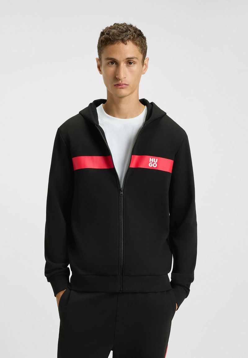 Felpa nera con zip realizzata in tessuto liscio, caratterizzata da una striscia orizzontale rossa brillante e un logo. Include un cappuccio con cordino e tasche laterali.