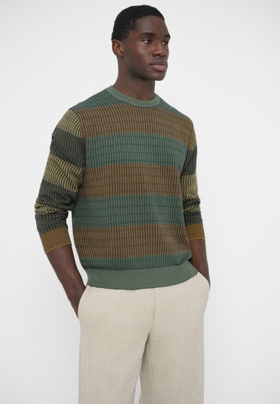 Zielony i brązowy sweter w wzory z prążkowaną fakturą i kontrastującym paskowym wzorem. Okrągły dekolt. Noszony z jasnymi spodniami.