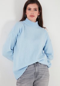 Gai zils, adīts turtlenecks džemperis ar ribinātu apkakli un apakšmalu, ar noliktām plecu šuvēm un brīvu piegriezumu, kopā ar pelēkiem džinsiem.