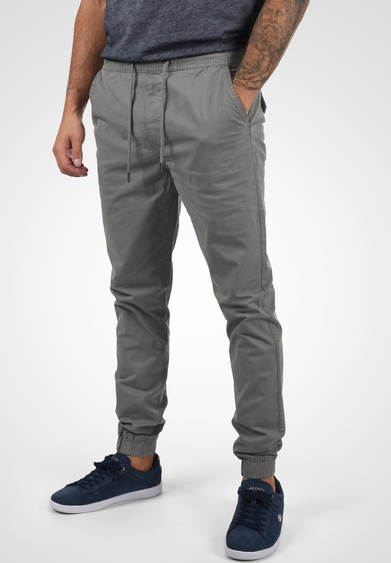 Gray, stretchy jogger pants with elastic waistband and cuffs. Features side pockets and a smooth texture. Worn with navy sneakers.