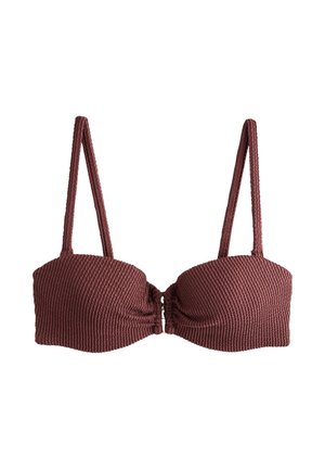 Femeie cu păr lung și închis la culoare, purtând un top de bikini mov închis, stă cu fața spre ocean, cu apă liniștită și cer senin în fundal.