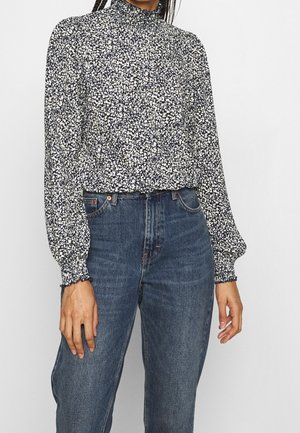 Vrouw draagt een zwarte en witte bloemenblouse met hoge hals en lange mouwen, gecombineerd met hoge taille blauwe spijkerbroek, staand tegen een effen achtergrond.