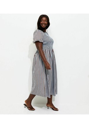 Kobieta idąca w niebiesko-białej sukience midi w paski z bufkowymi rękawami, nosząca czarne sandały na obcasie na jednolitym białym tle.