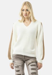 Witte V-hals gebreide trui met bruine gewatteerde mouwen. Heeft ribgebreide boorden en een casual pasvorm, gecombineerd met versleten grijze jeans.