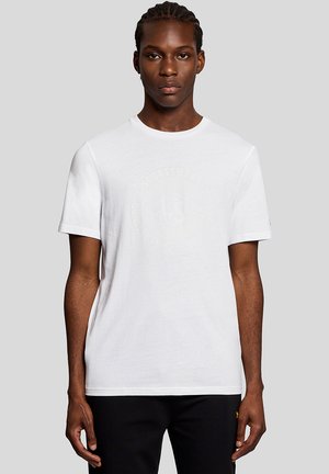 Ung man med flätat hår klädd i en enkel vit T-shirt med en svagt präglad cirkulär logotyp, stående framför en ljusgrå bakgrund.