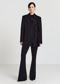 Blazer nero su misura con cintura da annodare in vita e rever appuntiti, abbinato a pantaloni neri a zampa, creando una silhouette definita e un look moderno.