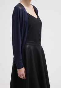 Námořnický otevřený kardigan s dlouhými rukávy, se zpěvnými rameny. Nosený přes černý hedvábný top a černou koženou sukni. Hladká textura.