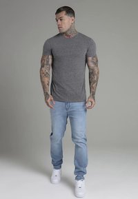 Pilka aptempta marškinėliai, derinami su šviesiai mėlynais džinsais ir baltais sportbačiais. Asmenybė turi ryškius tatuiruočius ir stilizuotus plaukus, stovi prieš neutralią foną.