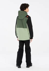 Grüne und schwarze Jacke mit Kapuze, die ein Farbblockdesign und elastische Bündchen aufweist. Getragen mit schwarzen Hosen und robustem Schuhwerk.