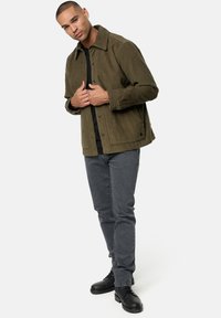 Olijfgroene corduroy jas met een kraag, knopen aan de voorkant en zijzakken, gecombineerd met grijze jeans en zwarte schoenen, staande pose.
