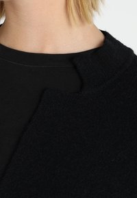 Svart ullrock med hög krage, med en strukturerad yta och en något asymmetrisk design, lagd över en svart stickad topp.