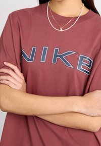 Person som bär en vinröd Nike-t-shirt med en stor marinblå och vit logotyp, med armarna i kors, samt lager av guld- och pärlhalsband med en hjärtformad berlock.