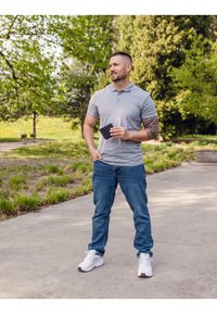 Gray polo shirt, blue jeans, and white athletic shoes. The person holds a black wallet, with visible arm tattoos and a natural outdoor setting.