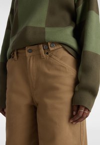 Maglione a fantasia verde e marrone abbinato a pantaloni marroni chiari con due bottoni argentati e dettagli di cucitura diagonale.