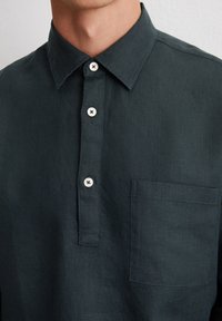 Chemise en lin vert foncé avec un col à boutons, dotée de trois boutons visibles, d'une poche poitrine et d'un tissu texturé.