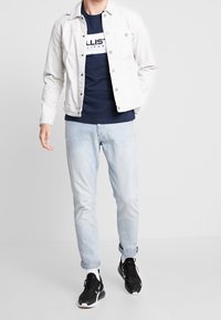 Vit denimjacka över en marinblå T-shirt, ljusblå jeans och svarta sneakers. Har knappar och en avslappnad passform.