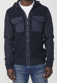 Marieblå zip-up hoodie med fleecepaneler, två bröstfickor, svarta dragkedjor och stickade mudd. Har en huva med dragsko och mjuk textur.