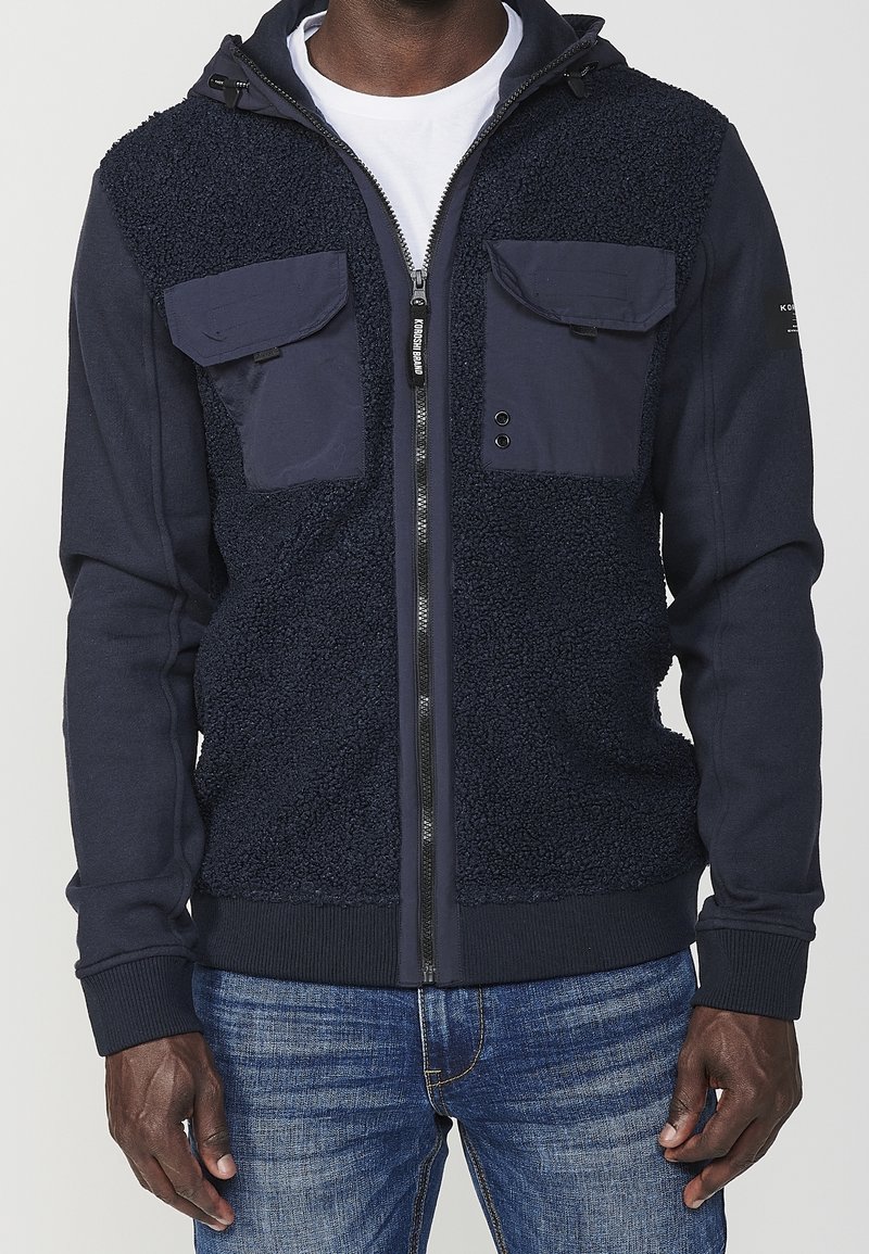 Marieblå zip-up hoodie med fleecepaneler, två bröstfickor, svarta dragkedjor och stickade mudd. Har en huva med dragsko och mjuk textur.