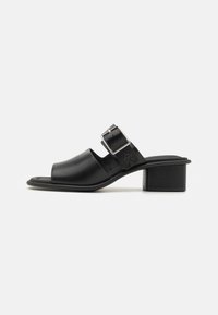 Sandale en cuir noir avec une large sangle sur le pied et un bout carré. Comprend une boucle en métal argenté et un petit talon carré. Semelle texturée.