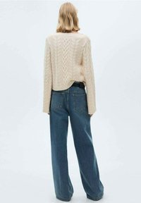 Pull en maille beige clair avec motif en câble ; associé à un jean bleu ample. Le pull a un ourlet arrondi et des manches larges.