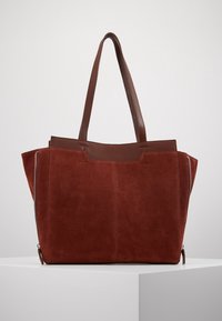 Grand sac fourre-tout en suède rouge avec des accents en cuir marron et de longues poignées ; présente des zips latéraux et une forme structurée.