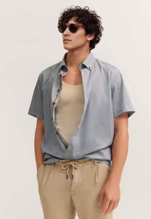 Mann mit lockigem Haar, der eine Sonnenbrille, ein gemustertes kurzärmeliges Hemd über einem beigen Tanktop und beige Kordelzughose trägt, die Hände in den Taschen.