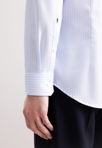 Witte overhemd met lichtblauwe verticale strepen, voorzien van een spitse manchet en een geborduurd zwart bloemenlogo op de mouw.