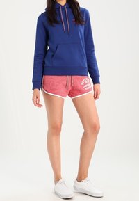 Felpa blu con tasca frontale e cordino, abbinata a pantaloni corti rossi con texture e dettagli bianchi, indossata con sneakers bianche.