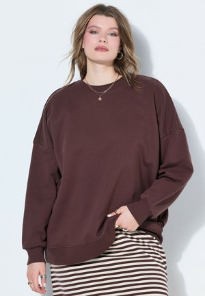 Młoda kobieta z włosami do ramion, ubrana w luźną brązową bluzę, pasiastą brązowo-białą spódnicę, złoty naszyjnik i kolczyki-koła.