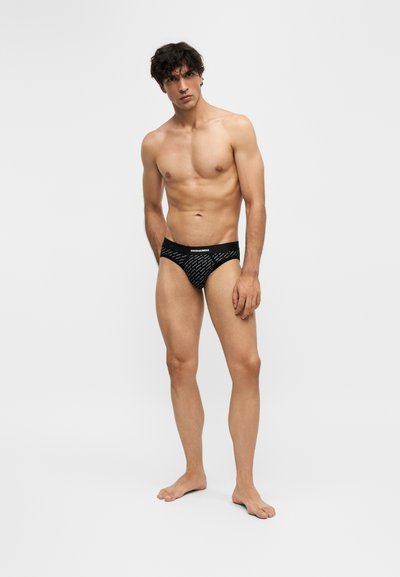 Jeune homme aux cheveux bouclés portant un slip noir à motifs, debout pieds nus sur un fond clair uni.
