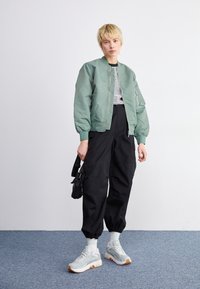 Vaaleanvihreä bomber-takki, raidallinen paita, mustat cargo-housut, harmaat lenkkarit vaaleilla yksityiskohdilla, pidetään pientä mustaa kassia. Rentoa katutyyliä.