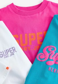 Tre t-shirts i pink, hvid og turkis med forskellige logoer. Den pink har orange skrifttype, den hvide har lilla, og den turkise har pink accenter.