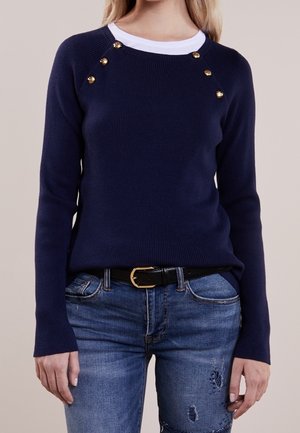 Nainen pukeutuneena tummansiniseen ribbineulepaitaan, jossa on kultaiset napit hartioiden lähellä, valkoisen paidan päällä ja parina kulutetut siniset farkut.