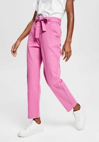 Pantaloni in denim rosa con vita annodata, tasche laterali e vestibilità rilassata, abbinati a sneakers bianche su uno sfondo neutro.