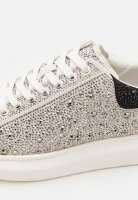 Baskets avec une tige blanche texturée ornée de clous en argent et de strass, complétées par une touche de noir et des lacets blancs.
