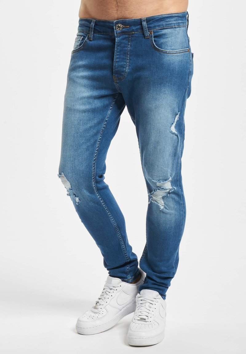 Homme portant un jean bleu slim déchiré et des baskets blanches, debout sur un fond blanc.