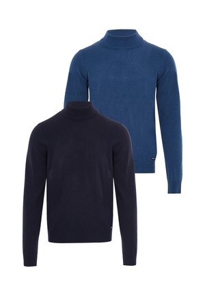 Homme portant un pull à col roulé bleu, un jean foncé et des baskets blanches, debout et regardant sur le côté sur un fond blanc.