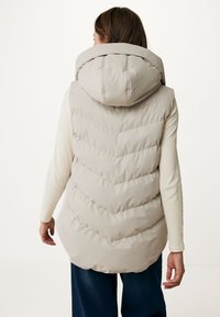 Gilet doudoune beige avec un col montant, présentant un motif en chevron, une texture lisse et un ourlet arrière long. Porté par-dessus un pull léger.