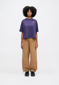 Una persona in piedi di fronte, indossa una maglietta viola a maniche corte, pantaloni marroni larghi e scarpe nere, su uno sfondo bianco semplice.