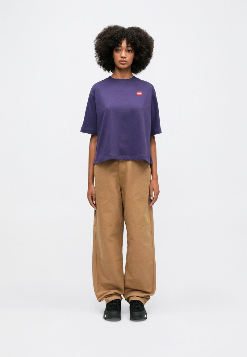 Una persona in piedi di fronte, indossa una maglietta viola a maniche corte, pantaloni marroni larghi e scarpe nere, su uno sfondo bianco semplice.