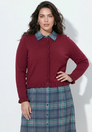 Vrouw met lang golvend haar draagt gouden oorringen, een bordeauxrode cardigan met gerimpelde randen en een blauwgroene geruite jurk met knopen.
