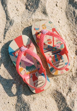 Tongs pour enfants avec des brides roses et des personnages de dessins animés colorés, reposant sur un sol sableux grossier au soleil.