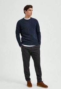 Νεαρός άντρας στέκεται με τα χέρια στις τσέπες, φορώντας ναυτικό πουλόβερ, λευκή μπλούζα από κάτω, σκούρους παντελόνες και καφέ παπούτσια, κοιτάζοντας στο πλάι.