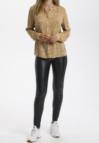 Senfgelbe Blume Langarmhemd mit Knöpfen, kombiniert mit glänzenden schwarzen Leggings und weißen Turnschuhen mit orangefarbenen Akzenten.