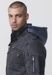 Grå denimjacka med en blå tyghuva, som har flera fickor med dragkedja och sydda detaljer på axlarna och ärmarna.