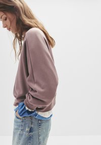 Felpa texturizzata in mauve con vestibilità rilassata e maniche arrotolate, indossata sopra una maglietta blu, abbinata a jeans chiari strappati.