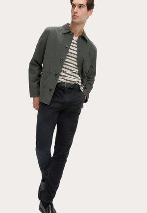 Olijfgroene jas met kraag, knoopsluiting en voorzakken, gedragen over een gestreept shirt. Zwarte broek en schoenen maken de outfit compleet.
