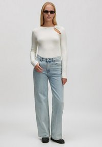 Weißer Langarm-Bodysuit mit einem Ausschnitt, kombiniert mit hellblauen weit geschnittenen Jeans und schwarzen spitz zulaufenden Schuhen.